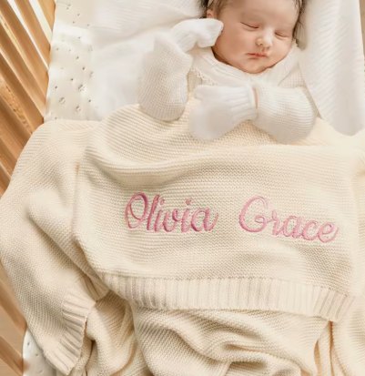 Newborn asleep under an apricot knit blanket embroidered with the name Olivia Grace in pink script